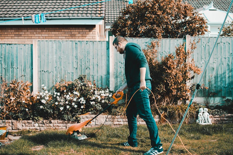 A woman and a man are working together in a well-maintained garden. The woman, wearing a white shirt, light blue jacket, and red gardening gloves, is holding a small potted evergreen shrub and a trowe